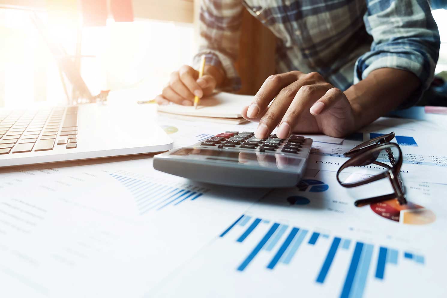 A man using a calculator to evaluate costs for his business and writing down the data on a notepad.